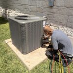 An HVAC technician kneeling next to a residential AC condenser, performing seasonal service for an HVAC maintenance plan.