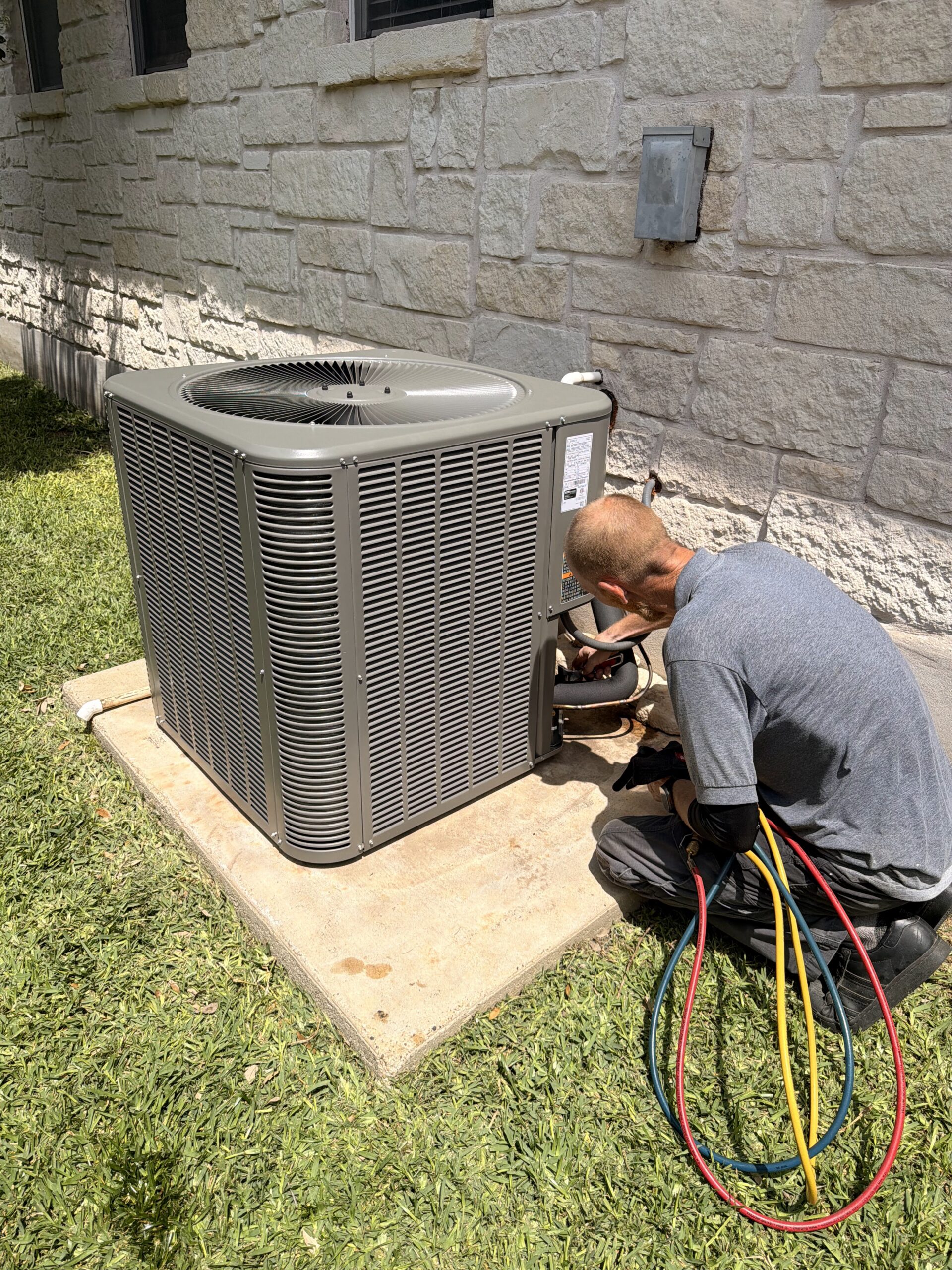 An HVAC technician kneeling next to a residential AC condenser, performing seasonal service for an HVAC maintenance plan.